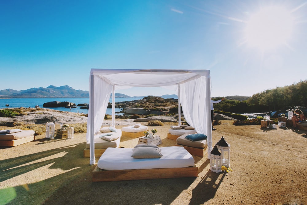 Un cadre romantique et élégant pour une cérémonie de mariage sur la plage, mettant en avant l'expertise de By Mademoiselle C en tant que wedding planner en Provence, Côte d'Azur et Corse. Ce lieu exceptionnel, avec ses vues imprenables sur la mer et les montagnes, est parfait pour une célébration grandiose. Les espaces de détente, ornés de lanternes et de coussins, créent une atmosphère intime pour un mariage sur mesure. Idéal pour un
