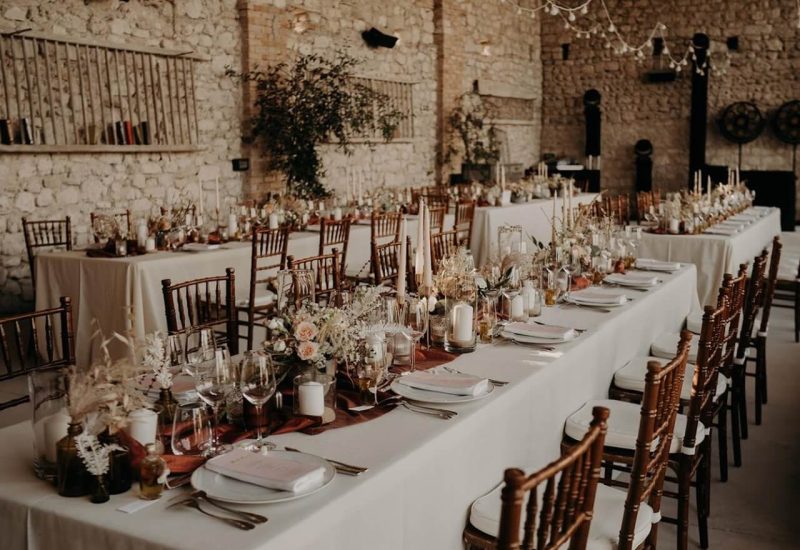An elegant stone reception hall, decorated for a luxurious celebration, showcasing By Mademoiselle C's expertise as a wedding planner in Provence, French Riviera &amp; Corsica. The tables, adorned with delicate floral arrangements and candles, create a romantic atmosphere for a bespoke wedding. This sophisticated setting, with its stone walls and warm ambience, is ideal for a large ceremony or a small wedding.