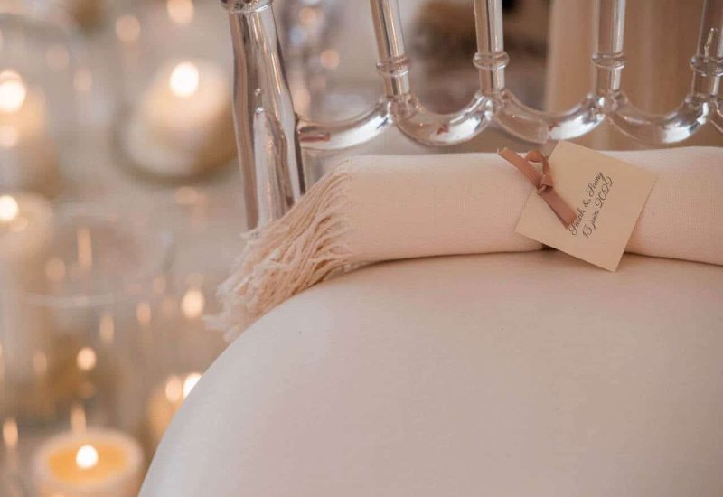 Un élégant détail de mariage mettant en avant un rouleau de serviette soigneusement présenté sur une chaise transparente, entouré de bougies scintillantes. Cette ambiance romantique est parfaite pour un grand événement organisé par By Mademoiselle C, le meilleur choix pour une planification de mariage de luxe en Provence, Côte d'Azur et Corse. Chaque élément est pensé pour créer une célébration sur mesure, faisant de chaque cérémonie un mariage de rêve dans des lieux d'exception. Que ce