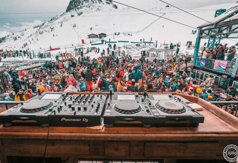 Une ambiance festive et dynamique au cœur des montagnes, où des milliers de personnes se rassemblent pour célébrer un événement unique. L'image montre des équipements de DJ sur une table en bois, avec une foule colorée en arrière-plan, évoquant une atmosphère de grand événement. Bien que cela ne soit pas une scène de mariage, l'énergie et l'excitation rappellent le caractère exceptionnel des célébrations. Folie Douce