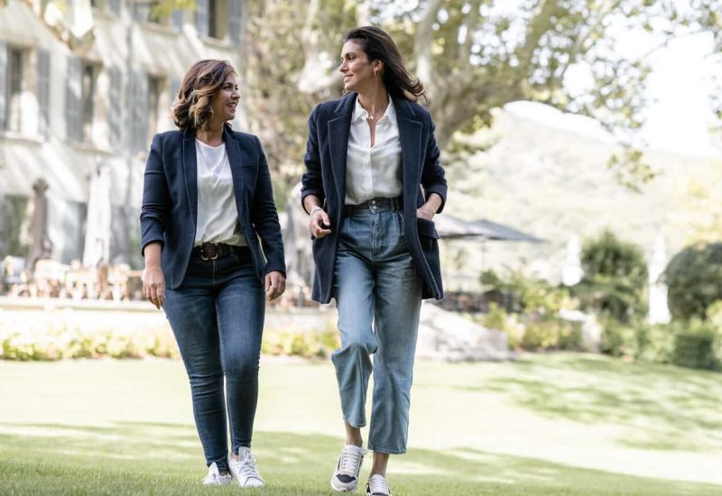 Two elegant wedding planners walk through a verdant garden, representing the expertise of 'By Mademoiselle C' as Wedding Planner in Provence, French Riviera &amp; Corsica. With a romantic and sophisticated setting, they embody the creation of luxurious, made-to-measure ceremonies. Their refined outfits reflect an upscale lifestyle, perfect for a grand event on an exceptional private estate.
