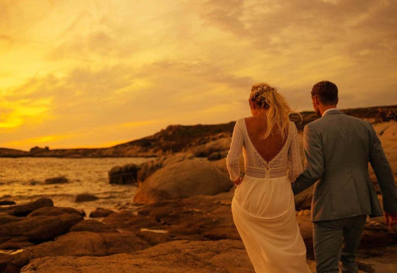 Romantic sunset wedding on the coast, featuring a couple walking hand-in-hand on rocks, illustrating By Mademoiselle C's expertise as a luxury wedding planner in Provence, French Riviera and Corsica. This unique moment captures the essence of a grand ceremony in an idyllic setting, perfect for a bespoke celebration. The warm colors of the sky add an elegant atmosphere and a touch of class.