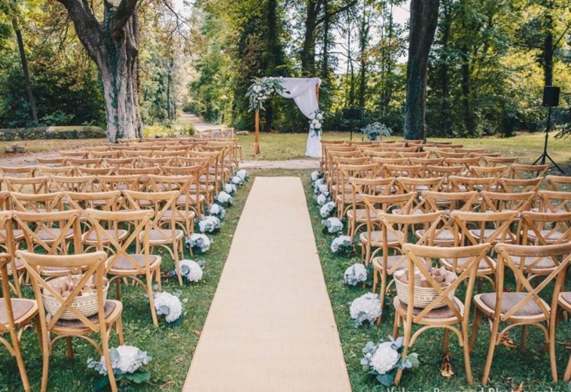 Romantic outdoor wedding ceremony, organized by By Mademoiselle C, showcasing an elegant, natural setting. Delicately arranged wooden chairs surround a carpeted aisle, creating a sophisticated atmosphere for a grand event. Ideal for a bespoke wedding on a private estate, this exceptional venue in Provence offers the perfect setting for a grand celebration. White flowers add a touch of charm to this