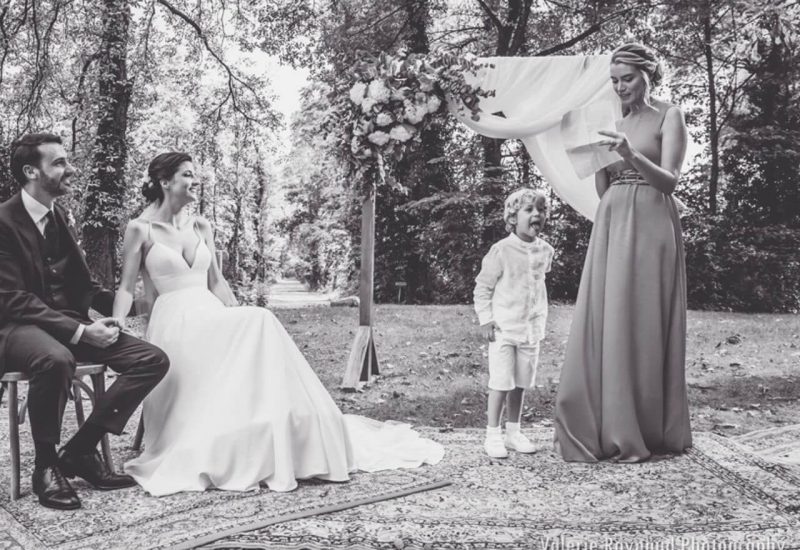Cérémonie de mariage romantique en plein air, mettant en scène un couple élégant assis dans un cadre naturel enchanteur, entouré de verdure. La mariée, vêtue d'une robe de luxe, et le marié, en costume raffiné, affichent une complicité touchante. Un enfant espiègle ajoute une touche de joie à cet événement sur mesure, organisé par By Mademoiselle C, spécialiste des mariages uniques en Provence, Côte d