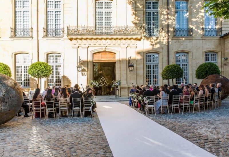 Luxurious outdoor wedding ceremony in an elegant setting, showcasing a private estate in Provence. The white driveway leads to a magnificent façade, surrounded by carefully tended greenery, creating a romantic atmosphere for a bespoke celebration. The elegantly dressed guests witness a unique event, a testament to By Mademoiselle C's expertise in organizing exceptional weddings. This scene illustrates