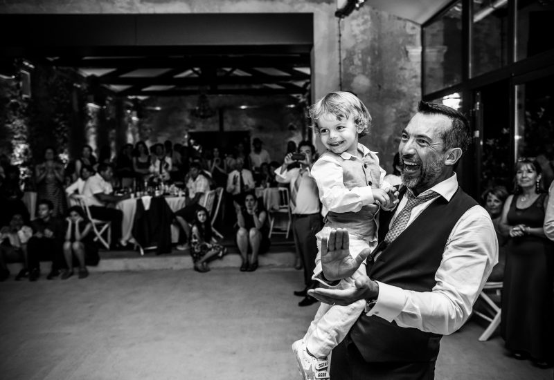 A grand celebration in an elegant setting, illustrating the expertise of 'By Mademoiselle C' as Wedding Planner in Provence, French Riviera &amp; Corsica. This image captures a joyous moment at a romantic wedding party, where a child is carried by a smiling man, surrounded by happy guests. The friendly, sophisticated atmosphere highlights the tailor-made organization of a unique event. This grand event, in