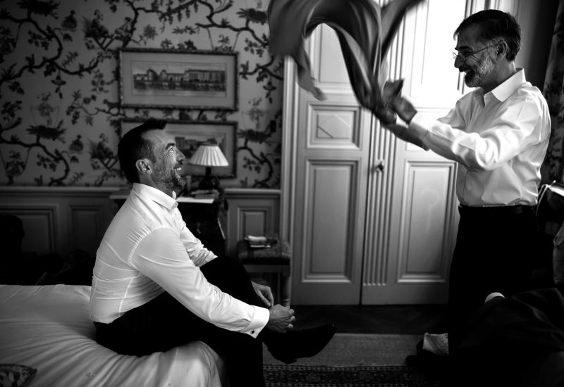 Un homme souriant s'apprête pour son mariage dans une chambre élégante, illustrant l'atmosphère romantique d'un grand événement. Cette scène, emblématique d'un mariage de luxe en Provence, Côte d'Azur et Corse, reflète l'organisation sur mesure de 'By Mademoiselle C'. La préparation minutieuse pour une cérémonie exceptionnelle souligne l'importance d'un service de planification de mariage sur mesure. L'élégance de la pièce, avec ses détails raffinés