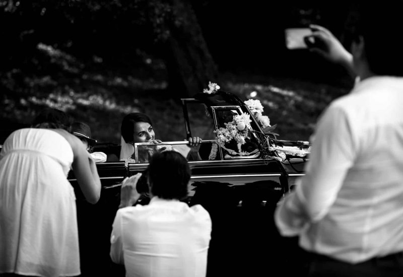 Une mariée souriante dans une voiture élégante, entourée de proches capturant des moments précieux, symbolise une célébration romantique et unique. Avec 'By Mademoiselle C', votre wedding planner en Provence, Côte d'Azur et Corse, chaque détail de votre mariage sur mesure est soigneusement organisé. Ce grand événement, dans un cadre enchanteur, évoque l’atmosphère sophistiquée d'une cérémonie de luxe, où des lieux exceptionnels comme des domaines privés