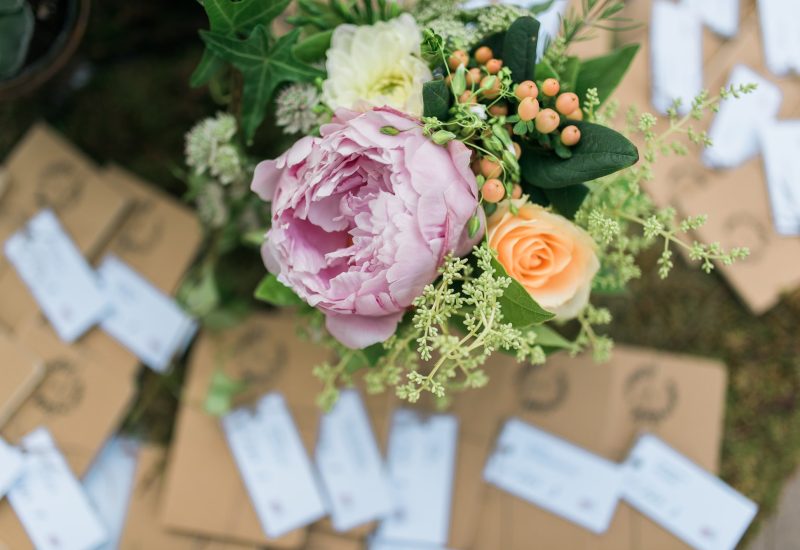 An elegant bouquet of pink peonies, orange roses and verdant foliage, symbolizing a romantic, luxurious wedding in Provence. Invitations carefully arranged in the background evoke a bespoke celebration, perfect for a grand event on a private estate. This enchanting setting highlights By Mademoiselle C's expertise in organizing exceptional ceremonies, offering a sophisticated ambience for a dream wedding on the Côte d'Azur.