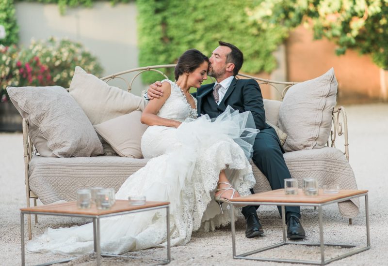 An elegant couple relax on a refined sofa in a romantic garden, illustrating a luxury wedding ceremony organized by By Mademoiselle C. This scene evokes a grand celebration in exceptional locations in Provence, the French Riviera and Corsica. With bespoke decorations and a sophisticated atmosphere, this event on a private estate promises a tailor-made wedding, combining romance and elegance to create an unforgettable dream.