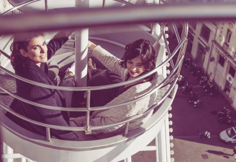 Deux femmes souriantes profitent d'une vue panoramique depuis une structure moderne, symbolisant l'élégance et la sophistication des événements organisés par 'By Mademoiselle C'. Cette image évoque l'atmosphère d'une planification de mariage sur mesure, idéale pour un mariage romantique ou une grande célébration dans des lieux d'exception en Provence, Côte d'Azur et Corse. L'expérience de luxe se reflète dans chaque détail.