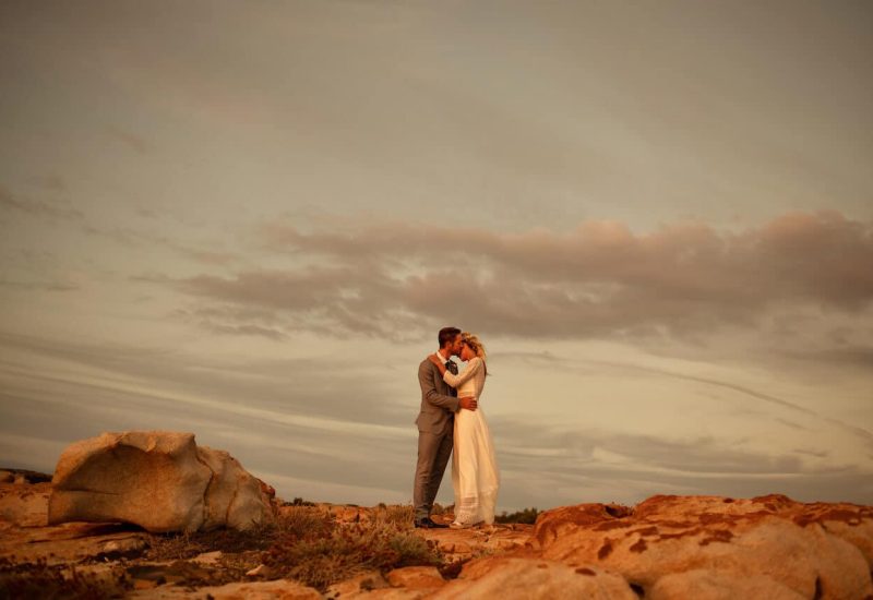 Loving couple kissing on a beach in Provence, creating a romantic and elegant ambiance. This luxury wedding ceremony, organized by By Mademoiselle C, embodies the essence of a bespoke event in exceptional surroundings. The natural setting, with its rocks and golden sky, evokes a grand celebration, perfect for a dream wedding in the French Riviera. Every detail has been carefully thought out to offer an experience