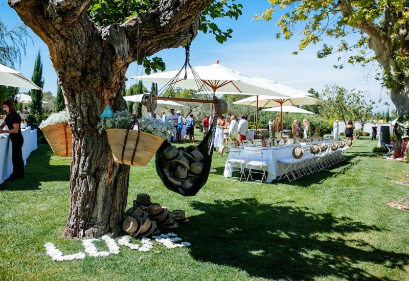 A luxurious wedding in an idyllic setting in the heart of a verdant garden in Provence, the French Riviera and Corsica. This romantic celebration features tailor-made organization, with a table elegantly set under parasols, surrounded by relaxed guests. Refined details, such as hats on the floor and delicate flowers, create a sophisticated atmosphere for a unique event. By Mademoiselle C
