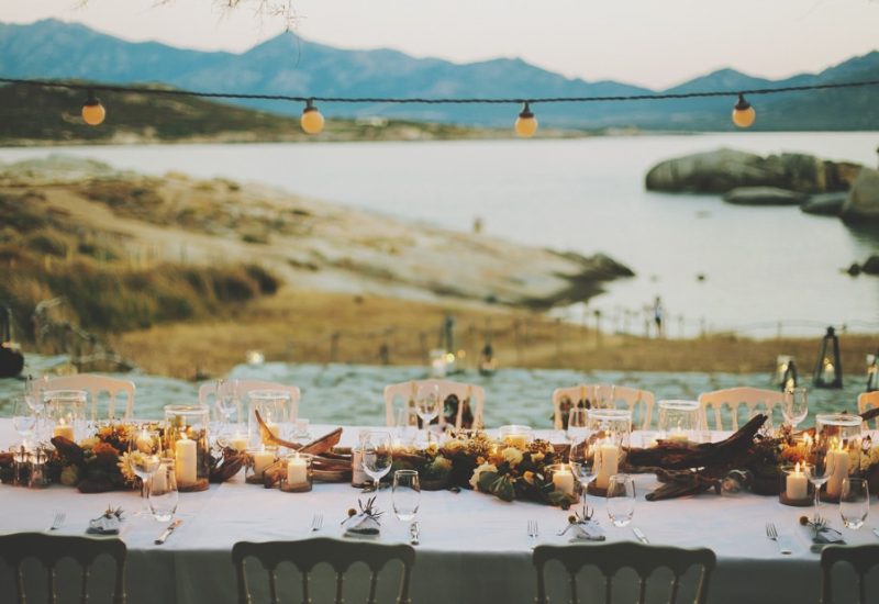 A sumptuous reception table for a romantic seaside wedding, organized by By Mademoiselle C, luxury wedding planner in Provence, French Riviera and Corsica. This idyllic scene features an elegant setting with twinkling candles and delicate flowers, creating an intimate atmosphere for a unique celebration. The natural setting, surrounded by mountains and rocks, underlines the exceptional character of this private estate.