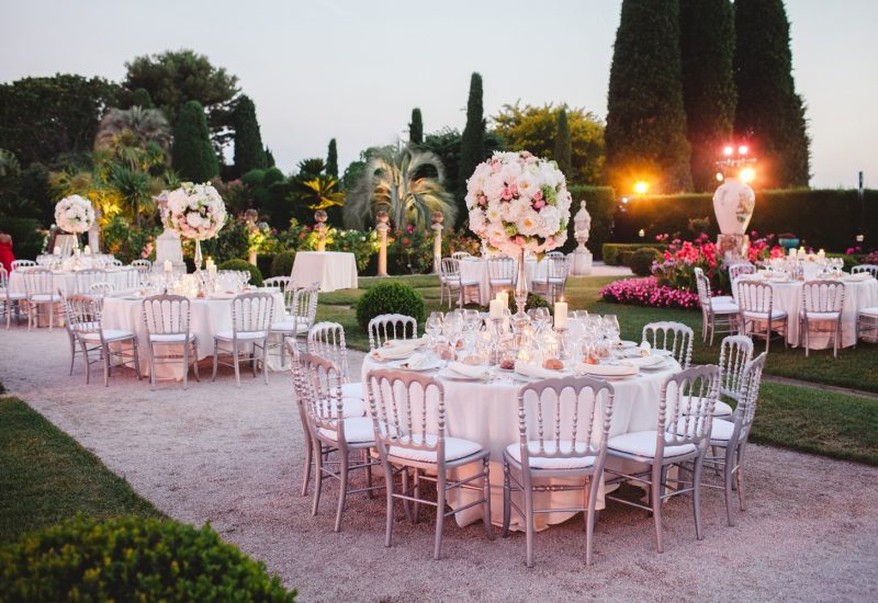 Une élégante réception de mariage en plein air dans un jardin luxuriant, mettant en valeur la beauté des lieux d'exception en Provence, Côte d'Azur et Corse. Les tables, soigneusement dressées pour un grand événement, sont ornées de superbes compositions florales, créant une atmosphère romantique et sophistiquée. Ce cadre idéal pour une célébration grandiose est parfaitement organisé par By Mademoiselle C, spécialiste des mariages sur mesure.
