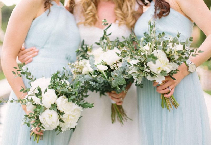 Trois demoiselles d'honneur portant des robes élégantes bleu pâle, entourées de bouquets de fleurs blanches et verdoyantes, incarnent l'atmosphère romantique d'un mariage de luxe. Cette image évoque l'organisation sur mesure d'une célébration grandiose, mettant en valeur le savoir-faire de By Mademoiselle C en tant que wedding planner en Provence, Côte d'Azur et Corse.