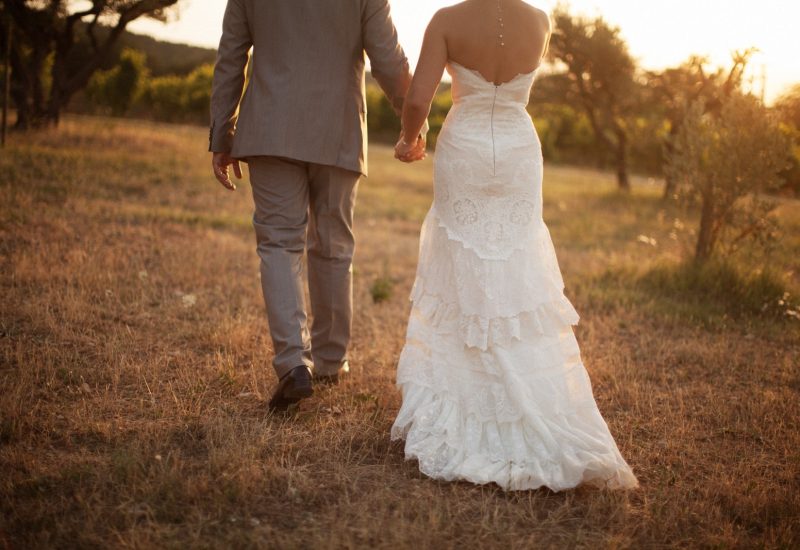 Un couple élégant marchant main dans la main dans un champ doré, symbolisant un mariage romantique et sur mesure. Ce moment capturé évoque l'atmosphère intime d'une célébration grandiose dans un domaine privé en Provence. La robe de mariée raffinée et le costume chic reflètent le style sophistiqué des événements organisés par 'By Mademoiselle C', experts en planification de mariages de luxe sur la Côte d'Azur et en Corse.