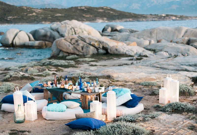 A romantic celebration by the sea, featuring an elegant dinner on soft cushions, ideal for a luxury wedding in Corsica. This exceptional setting, surrounded by coastal scenery and majestic mountains, embodies the essence of a bespoke wedding. With lanterns creating an intimate atmosphere and colorful bottles adding a touch of sophistication, this unique event promises an unforgettable experience for a grand wedding.