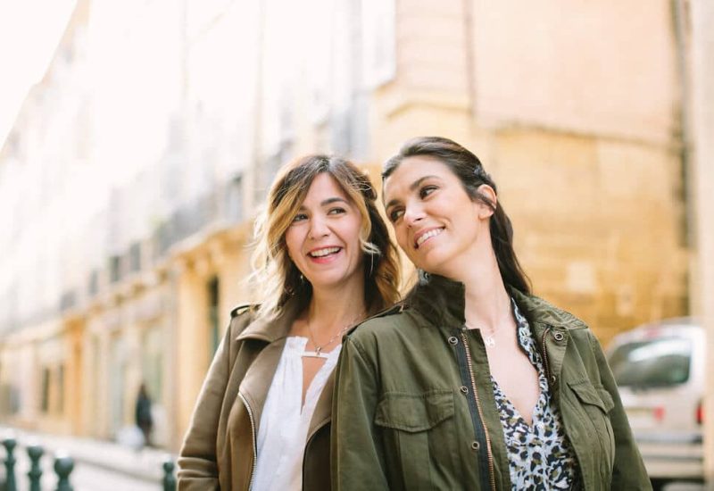 Two smiling women standing side by side on a picturesque street, symbolizing the elegance and sophistication of By Mademoiselle C, your expert in luxury wedding planning in Provence, French Riviera and Corsica. With a romantic background and soft colors, this image evokes the atmosphere of a grand celebration. Discover how our made-to-measure wedding services transform exceptional locations into private estates for