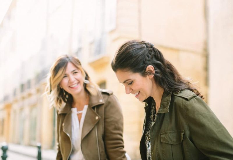 Two smiling women strolling down a picturesque street in Provence, representing the elegance and professionalism of 'By Mademoiselle C', a luxury wedding planning brand. As wedding planners in Provence, the French Riviera and Corsica, we are committed to organizing tailor-made events and grandiose ceremonies in exceptional locations. The scene evokes a romantic atmosphere, perfect for a grand celebration.