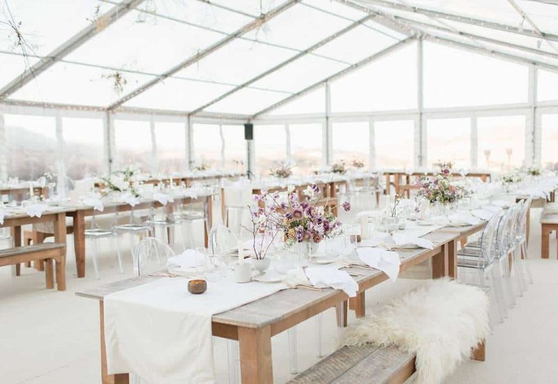 Une élégante salle de réception sous un chapiteau transparent, mettant en valeur une décoration raffinée pour une cérémonie romantique. Les tables en bois, soigneusement dressées, accueillent des fleurs délicates et des bougies, créant une atmosphère chaleureuse pour un grand événement. Ce cadre idyllique, situé dans un domaine privé, est parfait pour une célébration de mariage sur mesure. Avec des vues imprenables sur les vignobles environnants, cet espace incarne le