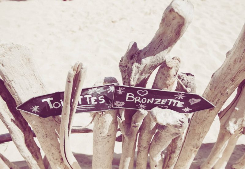 Un élégant panneau en bois sur une plage ensoleillée, indiquant les toilettes et une zone de bronzage, évoquant l'atmosphère décontractée d'un mariage romantique en bord de mer. 'By Mademoiselle C' se spécialise dans la création de célébrations grandioses et sur mesure, parfaites pour des événements uniques en Provence, Côte d'Azur et Corse. Ce cadre idyllique, avec le doux bruit des vagues, est l'endroit