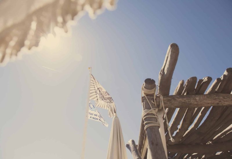 Drapeaux flottant au-dessus d'une élégante structure en bois sur une plage ensoleillée, évoquant une ambiance romantique et sophistiquée, idéale pour un grand mariage. Cette scène capture l'essence d'un événement sur mesure, parfait pour une célébration de luxe en Provence, Côte d'Azur et Corse. Avec des paysages enchanteurs en toile de fond, 'By Mademoiselle C' transforme chaque moment en un rêve inoubliable, offrant des lieux exception