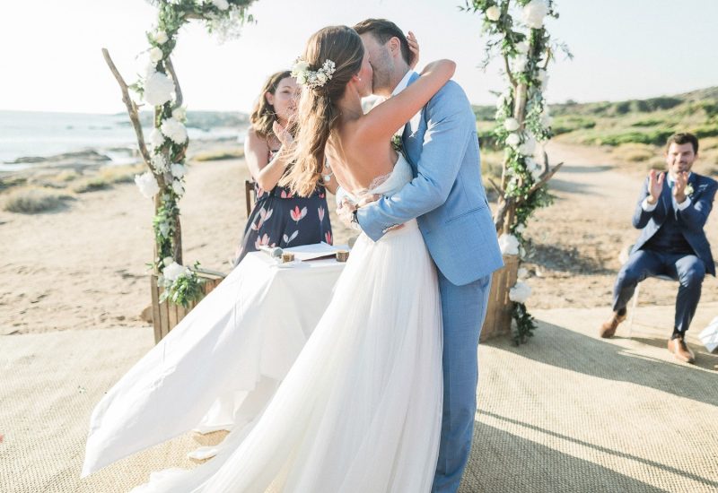 A romantic celebration of a luxury wedding on a picturesque beach, featuring a couple exchanging a kiss under a floral arch. The elegant, sophisticated atmosphere reflects By Mademoiselle C's expertise as a wedding planner in Provence, the French Riviera and Corsica. This bespoke event, held in an idyllic setting, evokes a unique wedding dream, perfect for a grand ceremony. Visit