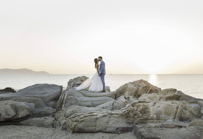 Romantic couple standing on rocks by the sea, capturing the essence of a luxury wedding in Provence, French Riviera and Corsica. This idyllic scene evokes a grand celebration, perfect for a bespoke event. The elegant, sophisticated atmosphere, with the sunset as a backdrop, makes this an exceptional private domain for a dream wedding. By Mademoiselle C excels in the