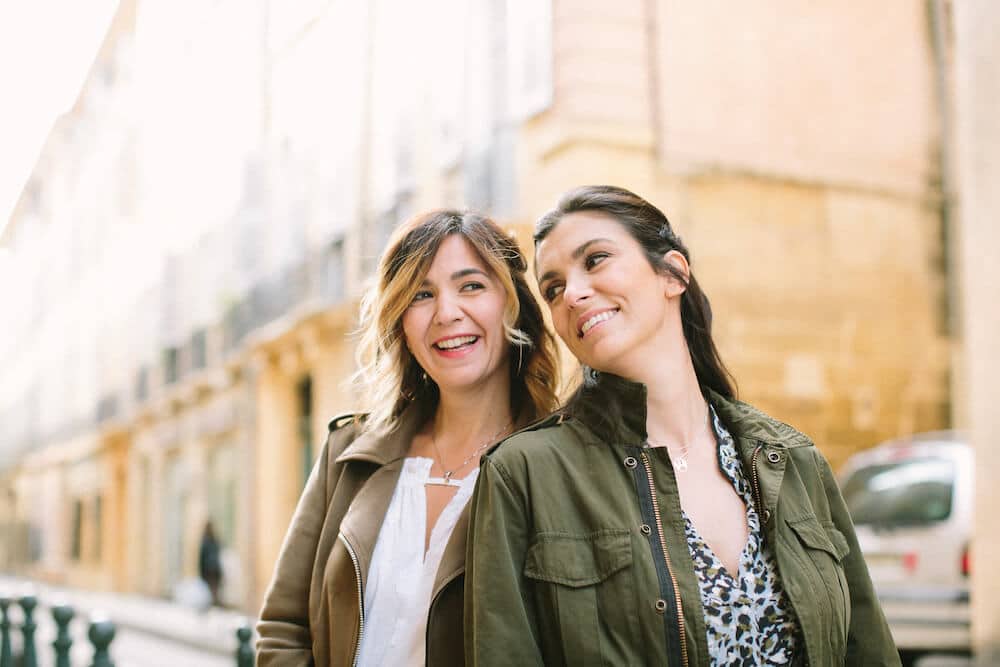 Deux femmes souriantes se tenant côte à côte dans une rue pittoresque, symbolisant l'élégance et la sophistication de By Mademoiselle C, votre experte en planification de mariage de luxe en Provence, Côte d'Azur et Corse. Avec un arrière-plan romantique et des couleurs douces, cette image évoque l'atmosphère d'une célébration grandiose. Découvrez comment nos services de mariage sur mesure transforment des lieux exceptionnels en domaines privés pour