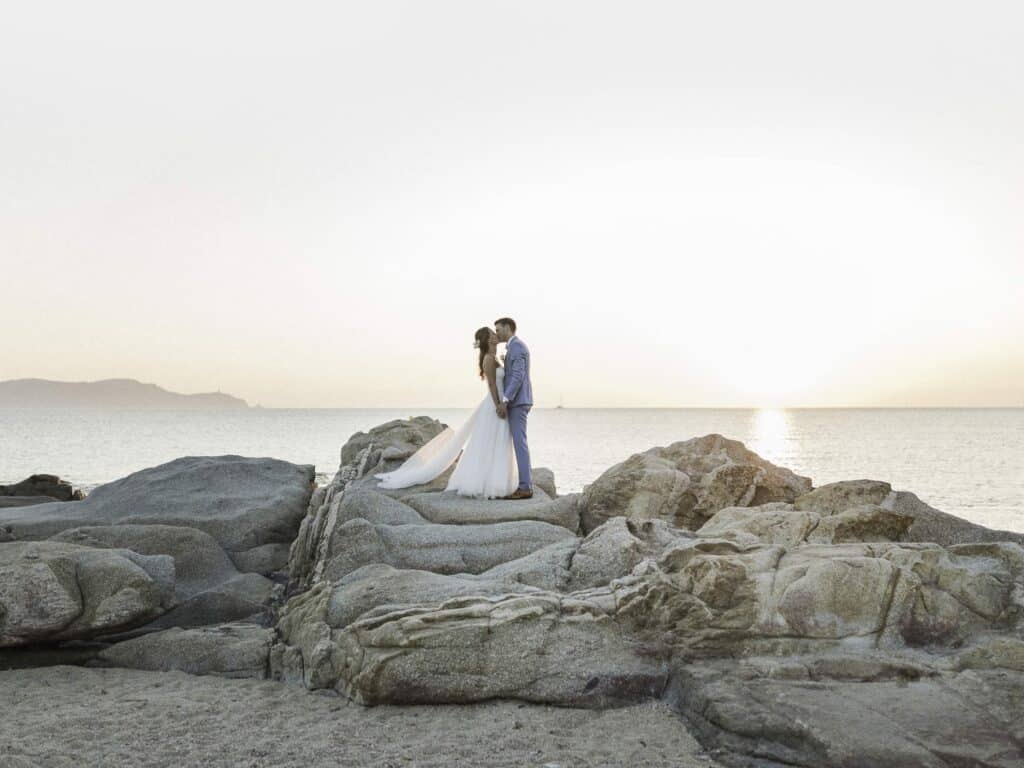 Un couple amoureux se tient sur des rochers au bord de la mer, au coucher du soleil, incarnant l'essence d'un mariage romantique en Provence. Ce moment magique illustre le savoir-faire de By Mademoiselle C en tant que planificateur de mariage de luxe, créant des célébrations sur mesure dans des lieux exceptionnels. Avec l'océan scintillant en arrière-plan, cette scène évoque une cérémonie grandiose, parfaite pour un événement unique dans un domaine privé