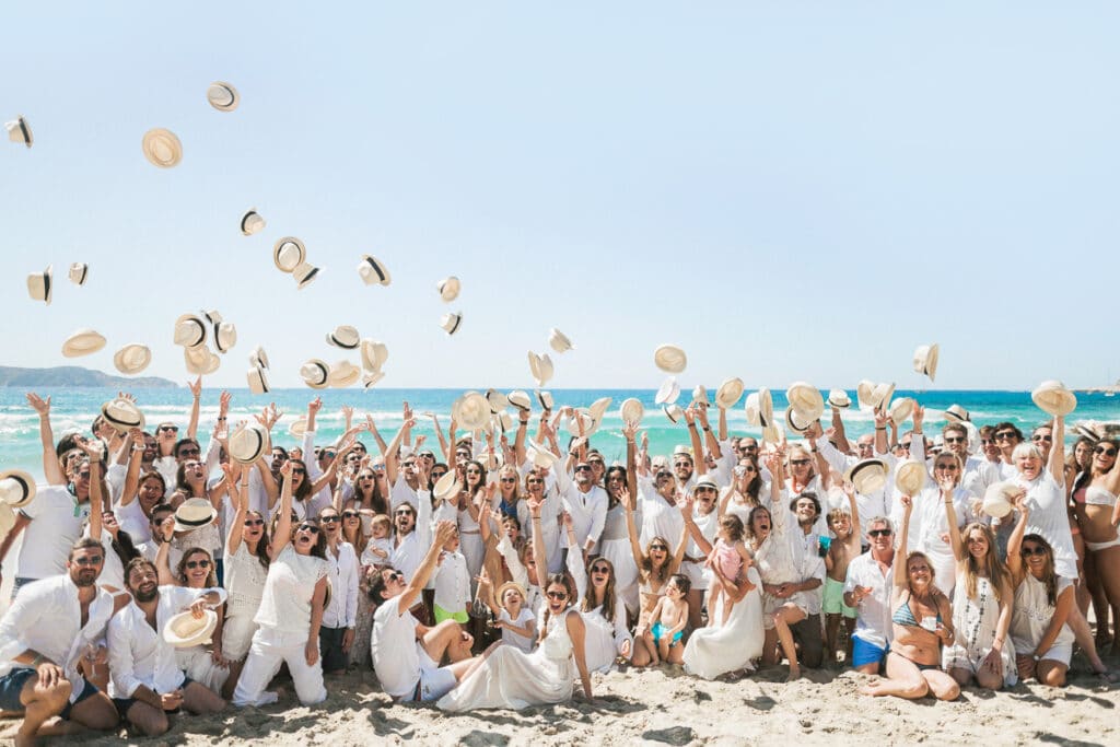 Une célébration en bord de mer réunissant des invités élégamment vêtus, illustrant un mariage romantique et une ambiance festive. Ce grand événement, organisé par 'By Mademoiselle C', incarne l'esprit d'un mariage de luxe en Provence, Côte d'Azur et Corse. Les invités, habillés de blanc, lancent des chapeaux dans les airs, créant une atmosphère joyeuse et mémorable. Ce moment époustouflant se déroule dans un
